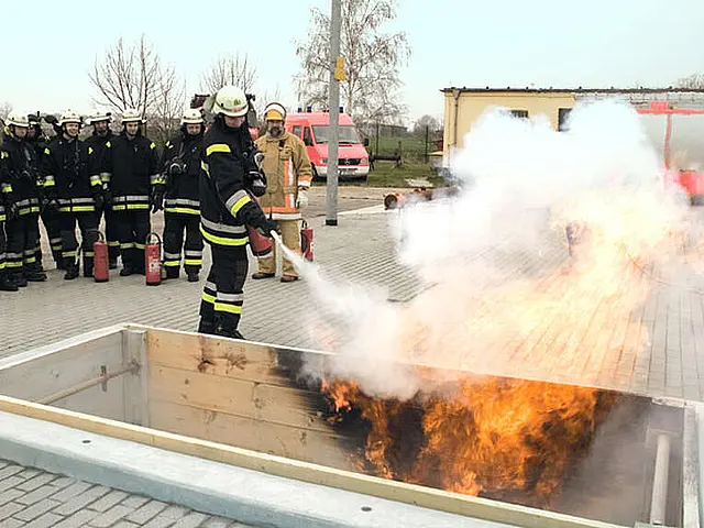 Feuerwehrleute üben den Einsatz von Feuerlöschern an einem brennenden Behälter, während eine Gruppe von Feuerwehrmännern zuschaut. Im Hintergrund sind Feuerwehrfahrzeuge zu sehen