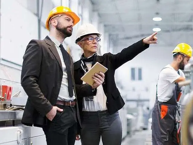 Ein Mann und eine Frau in Sicherheitshelmen besprechen etwas in einer Fabrikhalle, während sie auf einen Arbeiter im Hintergrund blicken. Der Mann hält einen Block in der Hand.