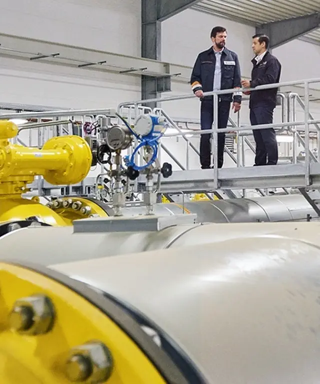 Industrieanlage mit großen gelben Gasarmaturen und Rohrleitungen in einer Halle; zwei Techniker stehen auf einer Wartungsbühne und sprechen miteinander.