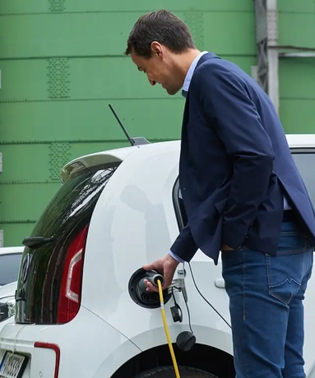 Mann lädt ein Elektroauto an einer Ladestation; im Hintergrund weitere parkende Autos vor einer grünen Industrieanlage.