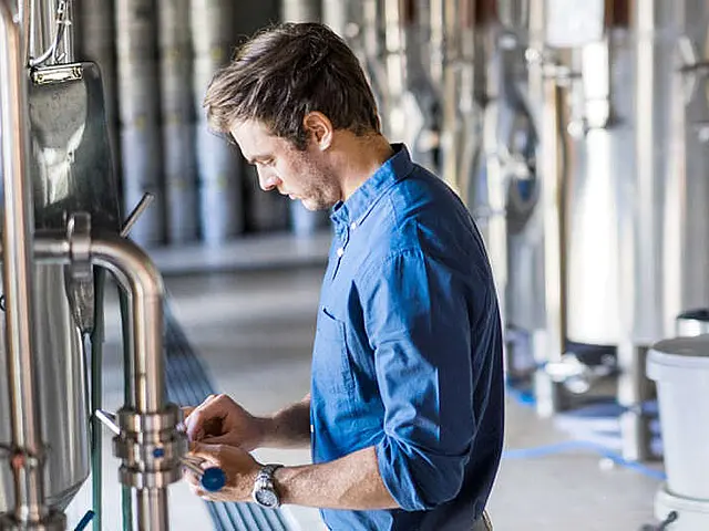Mann in blauem Hemd bedient ein Tablet in einer Industrieanlage mit Edelstahlrohren und technischen Anlagen.