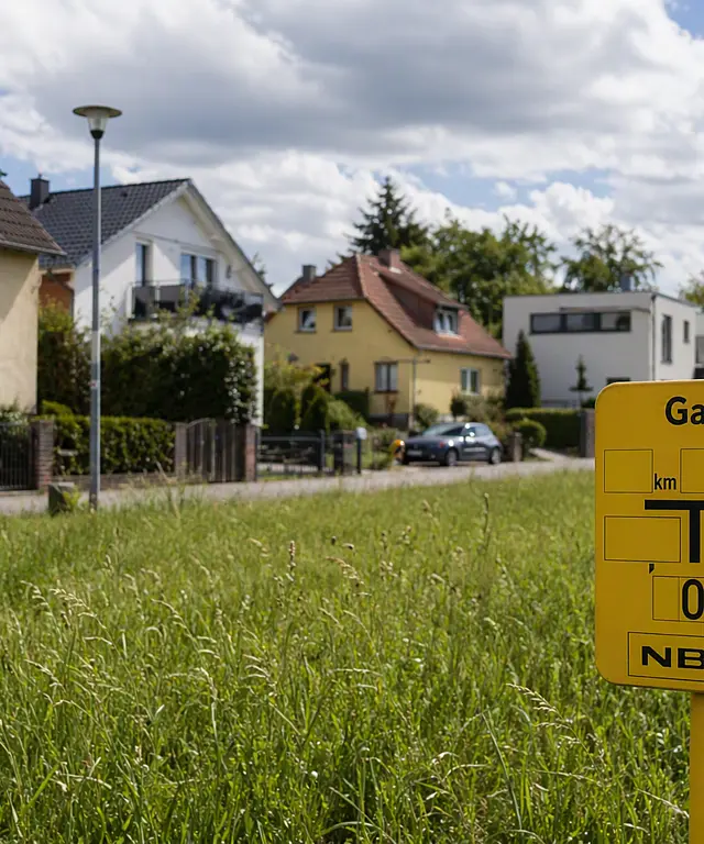 Gelbes Hinweisschild für eine Erdgasleitung auf einer Wiese vor einer Einfamilienhaussiedlung. Im Hintergrund sind Wohnhäuser, eine Straße und Bäume unter einem teilweise bewölkten Himmel zu sehen.