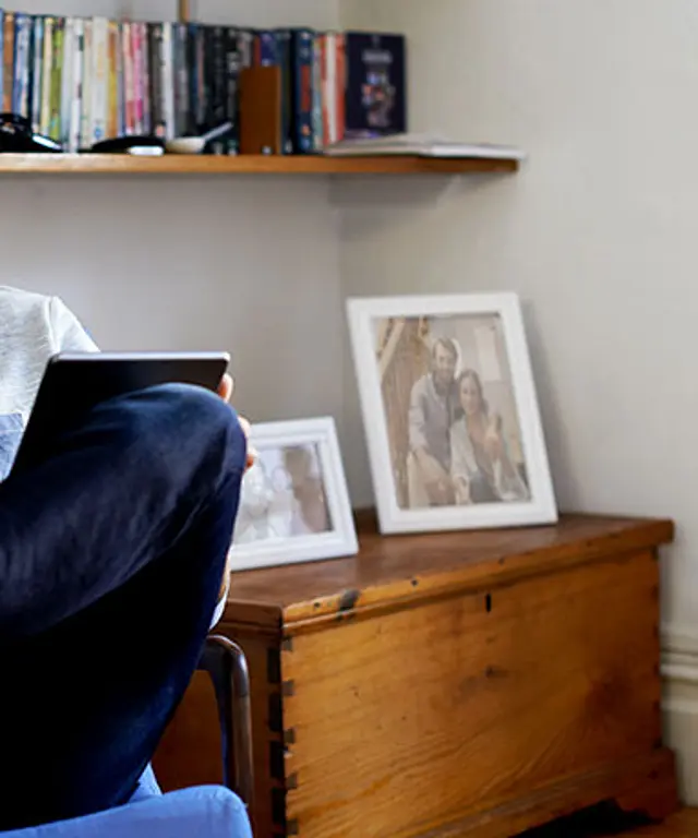 Mann sitzt entspannt in einem Sessel, trinkt aus einer Tasse und hält ein Tablet in der Hand. Im Hintergrund sind Bücher und Fotos auf einem Regal zu sehen