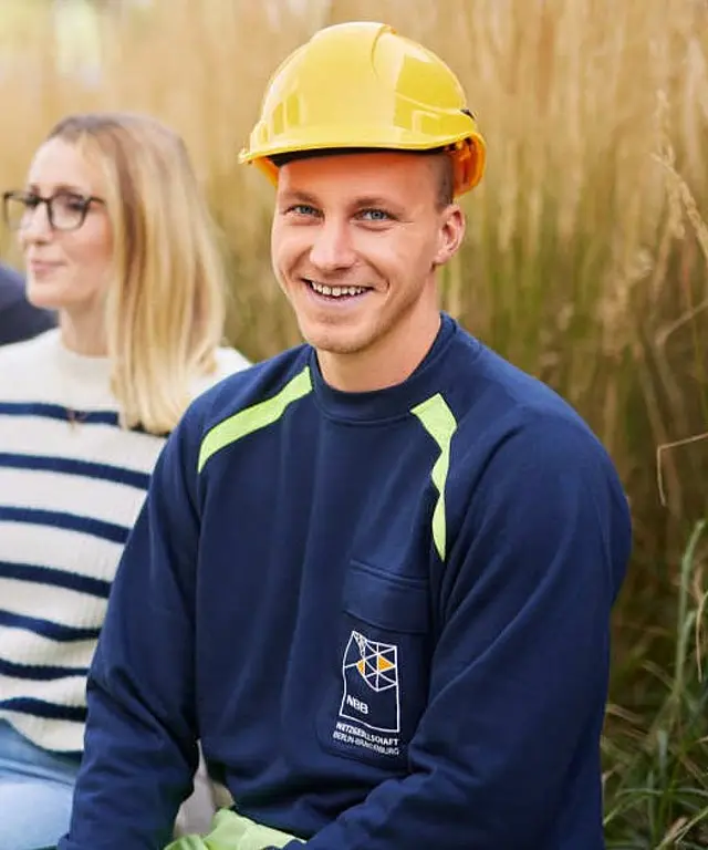 Drei Personen sitzen zusammen im Freien. Ein Mann trägt ein gelbes Sicherheitshelm und ein Arbeitsoutfit mit dem Logo 'NETZGESELLSCHAFT BERLIN-BRANDENBURG'. Zwei weitere Personen sitzen neben ihm und schauen in die Kamera